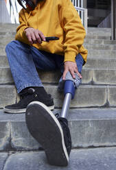 Behinderter Mann mit Beinprothese hält Smartphone, während er auf einer Treppe sitzt - VEGF03745
