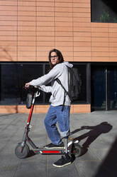 Young disabled man with electric push scooter standing on footpath during sunny day - VEGF03751