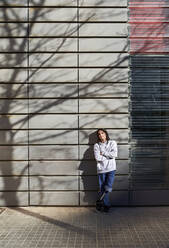 Young disabled man with arms crossed standing against wall with tree shadow - VEGF03770