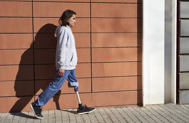 Junger Mann mit Beinprothese, der an einem sonnigen Tag an einer Mauer vorbeigeht - VEGF03775