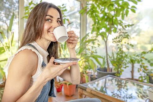 Smiling woman with gray eyes drinking coffee in sunroom - GGGF00948