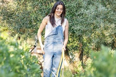 Smiling female with hand in pocket watering plants in backyard - GGGF00950