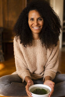 Smiling woman with herbal tea bowl sitting at home - AKLF00031