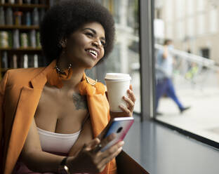 Glückliche Frau mit Smartphone und Kaffeetasse, die durch das Fenster eines Cafés schaut - JCCMF01167