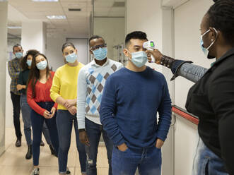 Group of multiracial colleagues in casual clothes and medical masks passing through procedure of body temperature control for coronavirus prevention before entering workplace - ADSF20563