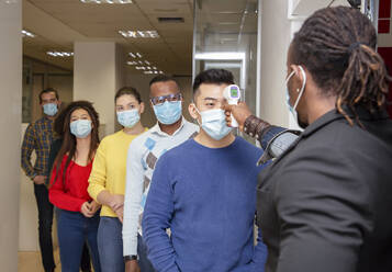 Group of multiracial colleagues in casual clothes and medical masks passing through procedure of body temperature control for coronavirus prevention before entering workplace - ADSF20564