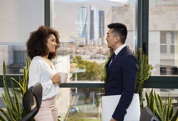 Seitenansicht einer positiven jungen afroamerikanischen Frau mit einer Tasse Kaffee und eines asiatischen männlichen Kollegen mit Laptop, die in der Nähe des Fensters stehen und über Neuigkeiten diskutieren, während sie zusammen in einem modernen Büro arbeiten - ADSF20565