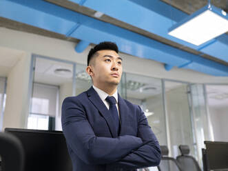 Serious young Asian male manager in formal suit looking away while standing near table with computers in contemporary coworking office - ADSF20571