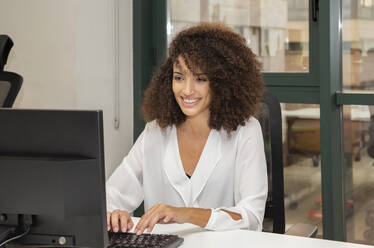 Young ethnic businesswoman with afro hairstyle sitting at table with computer while working on project in modern office - ADSF20577