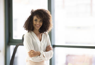 Cheerful young curly haired ethnic female in casual outfit standing near window and looking at camera while having break during work in modern workspace - ADSF20582