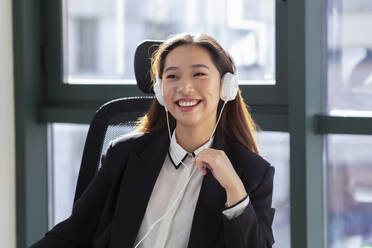 Glückliche junge asiatische Büroangestellte in formellem Outfit und Kopfhörern, die lachend ein Online-Gespräch in einem modernen Büro führt - ADSF20588