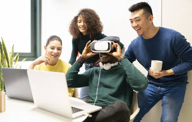 Unerkennbarer schwarzer Mann mit VR-Headset, der die virtuelle Welt erkundet, während er an einem Tisch mit Laptop sitzt, umgeben von fröhlichen multirassischen Kollegen an einem modernen Arbeitsplatz - ADSF20589