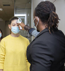 Frau in Freizeitkleidung und mit medizinischer Maske, die vor dem Betreten des Arbeitsplatzes das Verfahren zur Kontrolle der Körpertemperatur zur Vorbeugung gegen das Coronavirus durchläuft - ADSF20599