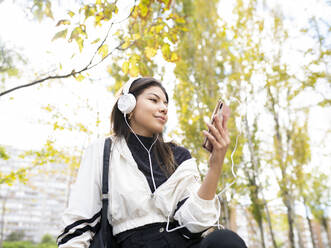 Von unten positiver junger ethnischer Frau in legerem Outfit, die mit Kopfhörern Musik hört und auf ihrem Handy surft, während sie im Park chillt - ADSF20602