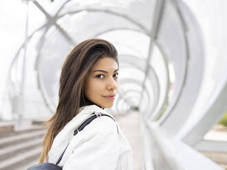 Seitenansicht einer einsamen, nachdenklichen jungen lateinamerikanischen Frau in trendigem Freizeitoutfit, die auf einer modernen Fußgängerbrücke steht und nachdenklich in die Kamera schaut - ADSF20603