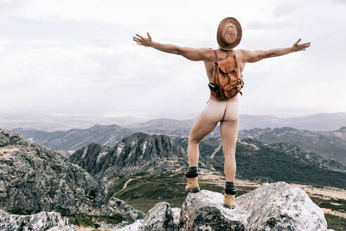 Back view of unrecognizable carefree male explorer with backpack and naked body standing on rock in mountains and enjoying freedom with outstretched arms - ADSF20618