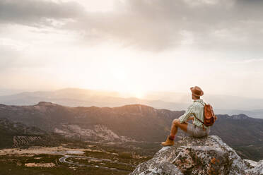 Seitenansicht von unerkennbaren reisenden Mann mit Rucksack sitzen auf einem Felsen im Hochland und bewundern erstaunlichen Sonnenuntergang über Bergkette - ADSF20619