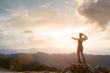 Rückenansicht eines unerkennbaren reisenden Mannes mit Rucksack, der auf einem Felsen im Hochland steht und einen fantastischen Sonnenuntergang über einer Bergkette bewundert - ADSF20621