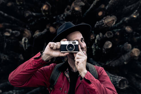 Zufriedener erwachsener Mann mit Hut, der ein Foto mit einer alten Fotokamera macht, während er an einem Stapel Holzstämme steht - ADSF20622