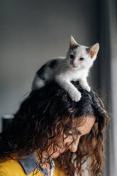 Seitenansicht der lächelnden Besitzerin mit weißem lustigen liebenswerten kleinen Kätzchen auf dem Kopf, das sich zu Hause ausruht - ADSF20632