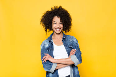 Positive junge lockig behaarte Afroamerikanerin in modischem Jeans-Outfit, die lächelt und in die Kamera schaut, während sie vor einem gelben Hintergrund steht - ADSF20635