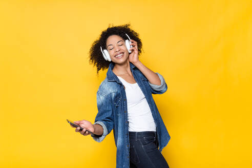 Fröhliche junge afroamerikanische Hipsterin im Jeanshemd, die Spaß hat und laute Musik über drahtlose Kopfhörer auf leuchtend gelbem Hintergrund genießt - ADSF20636