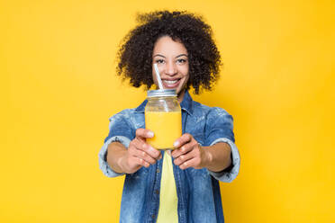 Fröhliche, junge, lockige, afroamerikanische Frau in Freizeitkleidung, die freundlich in die Kamera schaut und ein Glas mit gesundem, frischem Orangensaft zeigt, während sie vor einem gelben Hintergrund steht - ADSF20637