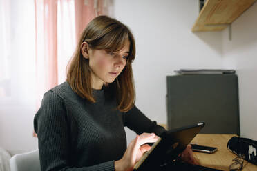 Blonde girl sitting at a desk at home looking at a tablet - ADSF20644