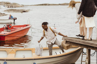 Smiling man helping female friends boarding boat by harbor - MASF21371