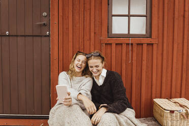 Female friends laughing while taking selfie on mobile phone against cottage - MASF21373