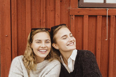 Happy female friends sitting side by side against cottage - MASF21376