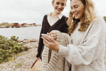 Smiling female friends using smart phone against sea during vacation - MASF21379
