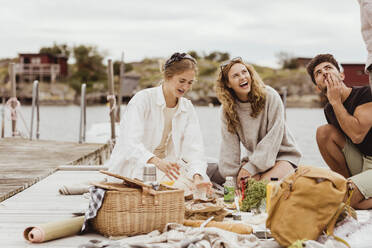 Friends laughing while having snacks during picnic at harbor - MASF21390