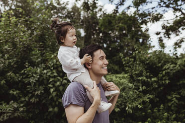 Happy father carrying baby son on shoulder by trees in park - MASF21394