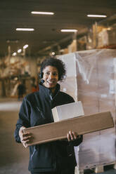 Lächelnde Geschäftsfrau mit Headset hält Pakete in einem Lagerhaus - MASF21453