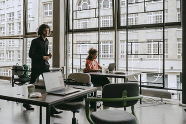 Female entrepreneur using laptop while male colleague walking by at coworking space - MASF21501