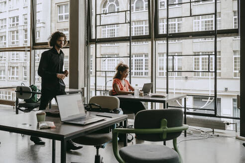 Unternehmerin benutzt Laptop, während ein männlicher Kollege im Coworking Space vorbeigeht - MASF21501