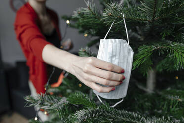 Die Hand einer Frau, die eine schützende Gesichtsmaske hält, hängt zu Hause am Weihnachtsbaum - OGF00864