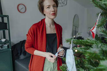 Lächelnde Frau vor dem Weihnachtsbaum im Wohnzimmer während der Pandemie - OGF00865