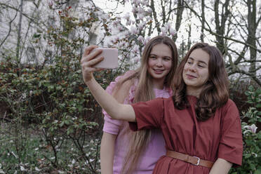 Smiling female friends taking selfie through mobile phone against magnolia tree - OGF00874