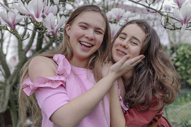 Fröhliche Freundinnen umarmen sich vor einem Magnolienbaum in einem öffentlichen Park - OGF00880