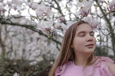 Teenage girl with eyes closed against magnolia tree in park - OGF00883