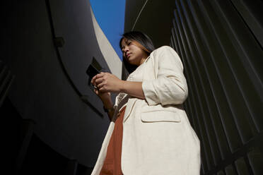 Young woman using smart phone while standing against gate during sunny day - VEGF03782