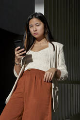 Confident young woman holding smart phone while standing against gate during sunny day - VEGF03790