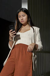 Selbstbewusste junge Frau, die ihr Smartphone hält, während sie an einem sonnigen Tag vor einem Tor steht - VEGF03790