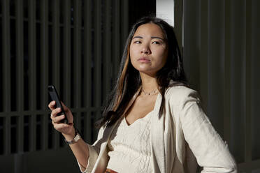 Nachdenkliche junge Frau mit Smartphone, die sich an einem sonnigen Tag an ein Tor lehnt - VEGF03791