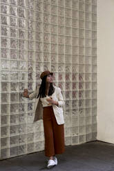 Young woman holding coffee cup looking away while standing against glass wall - VEGF03795