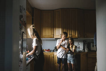 Sibling using smart phone while mother doing chores in kitchen - MASF21506