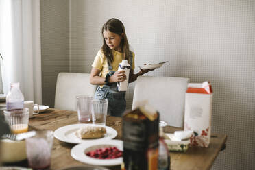 Vorpubertäres Mädchen mit Teller und Saftpackung am Esstisch stehend - MASF21558