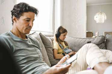 Vater benutzt sein Smartphone, während seine Tochter im Wohnzimmer mit dem Laptop e-Learning betreibt - MASF21588
